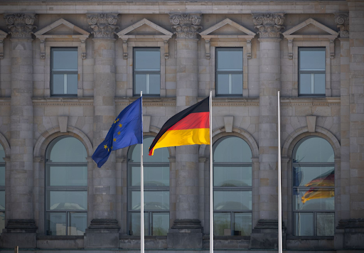 European Union and German flags flying side by side in front of a historic government building with stone columns and large arched windows.