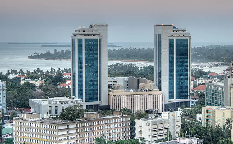 Aerial view of a modern city skyline featuring two tall glass office towers overlooking a coastal waterfront. Mid-rise commercial buildings, palm trees, and the shoreline are visible in the background, representing a major financial or business district in a coastal African city.