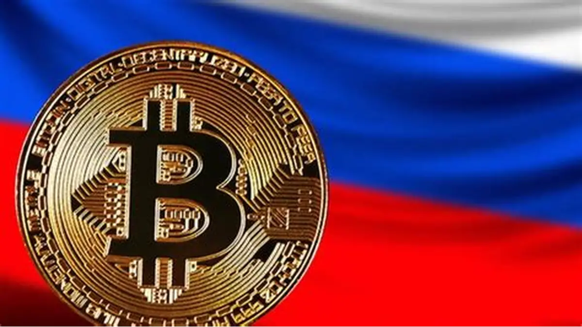 Gold Bitcoin coin displayed prominently in front of the Russian flag, featuring horizontal white, blue, and red stripes.