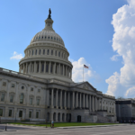 Trump to Meet With Senators Over Clarity Act on Thursday Front view of the United States Capitol building under a bright blue sky with large white clouds. And American flag is prominently visible, symbolizing the U.S. Congress, federal government, and national legislative policymaking.