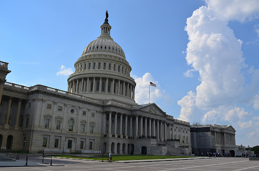 Front view of the United States Capitol building under a bright blue sky with large white clouds. And American flag is prominently visible, symbolizing the U.S. Congress, federal government, and national legislative policymaking.