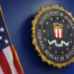 FBI Agent Accused of Stealing $1M in Crypto Allegedly Used ChatGPT for Vacation Plans The FBI seal displayed on a blue wall beside a partially visible American flag.