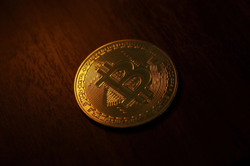 A gold Bitcoin coin resting on a dark wooden surface under warm lighting.