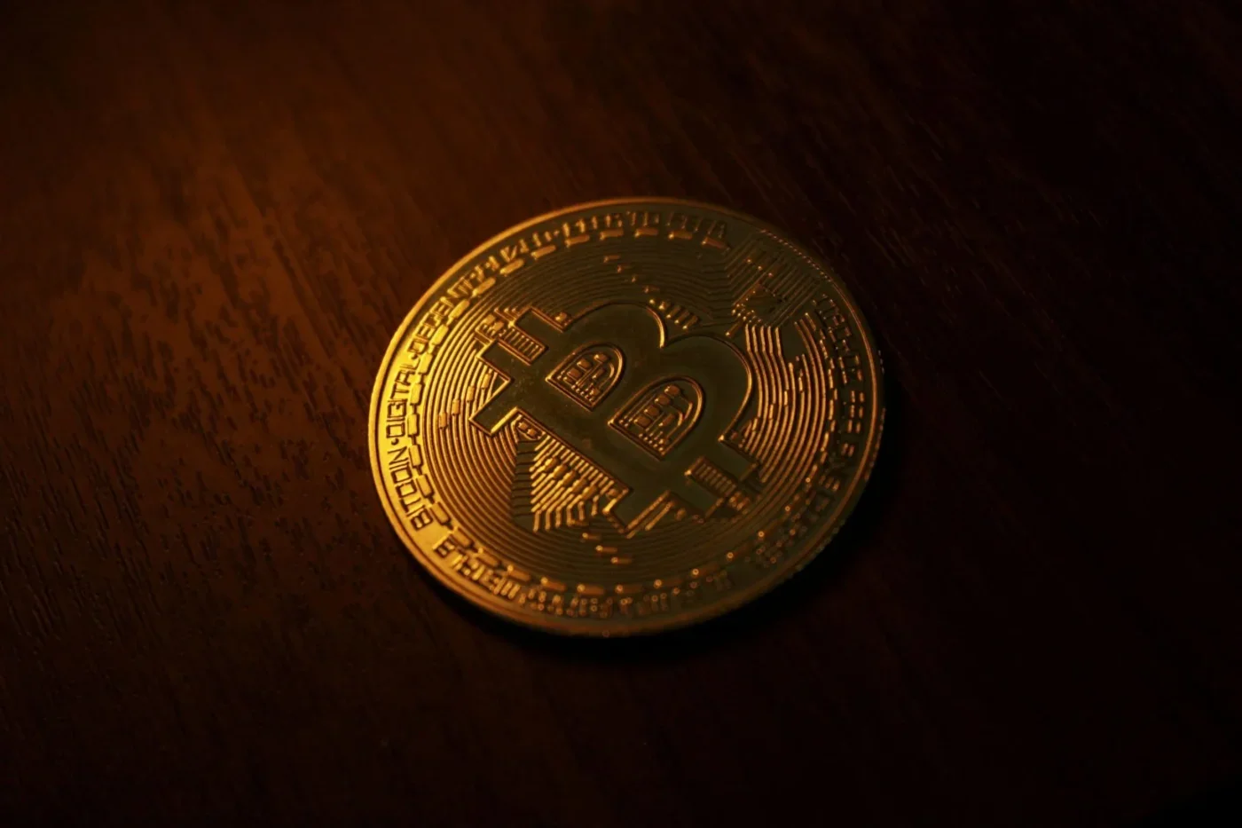 A gold Bitcoin coin resting on a dark wooden surface under warm lighting.