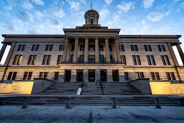 Front view of a historic neoclassical government building featuring a grand staircase, tall Corinthian columns, and a central domed tower beneath a blue sky with scattered clouds.
