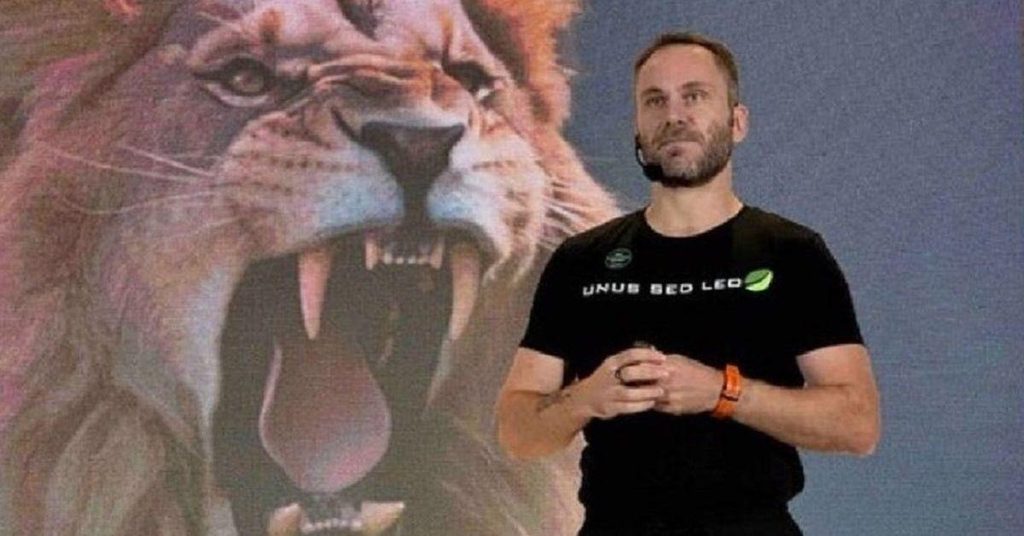 Image of Paolo Ardoino wearing a black shirt and an orange wristband with an image of a lion in the background